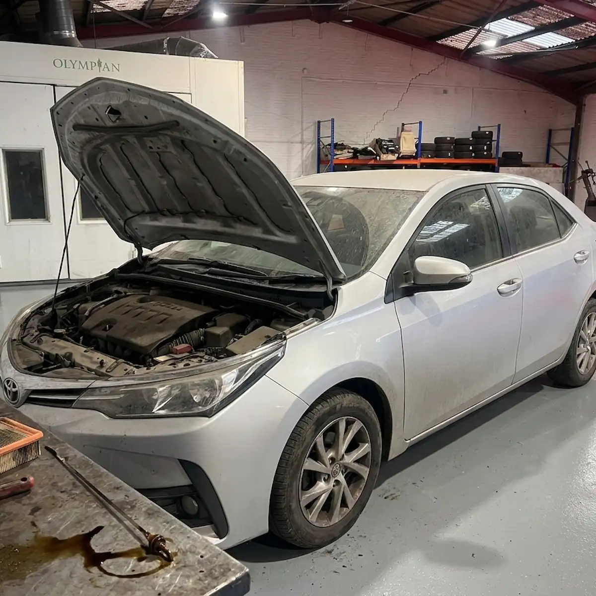 Dusty silver saloon with its bonnet raised in a workshop, a bench in front holding a dirty air filter, dipstick and tools.